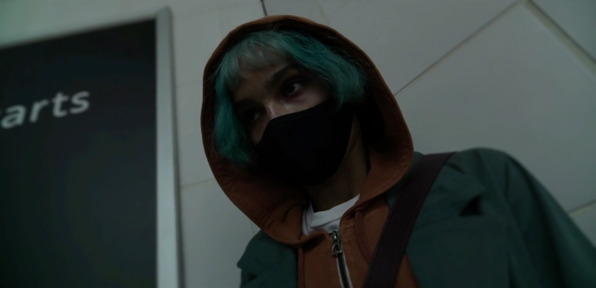 Angela (Zoe Kravitz) with her back to a train station wall, mask on, hood up.